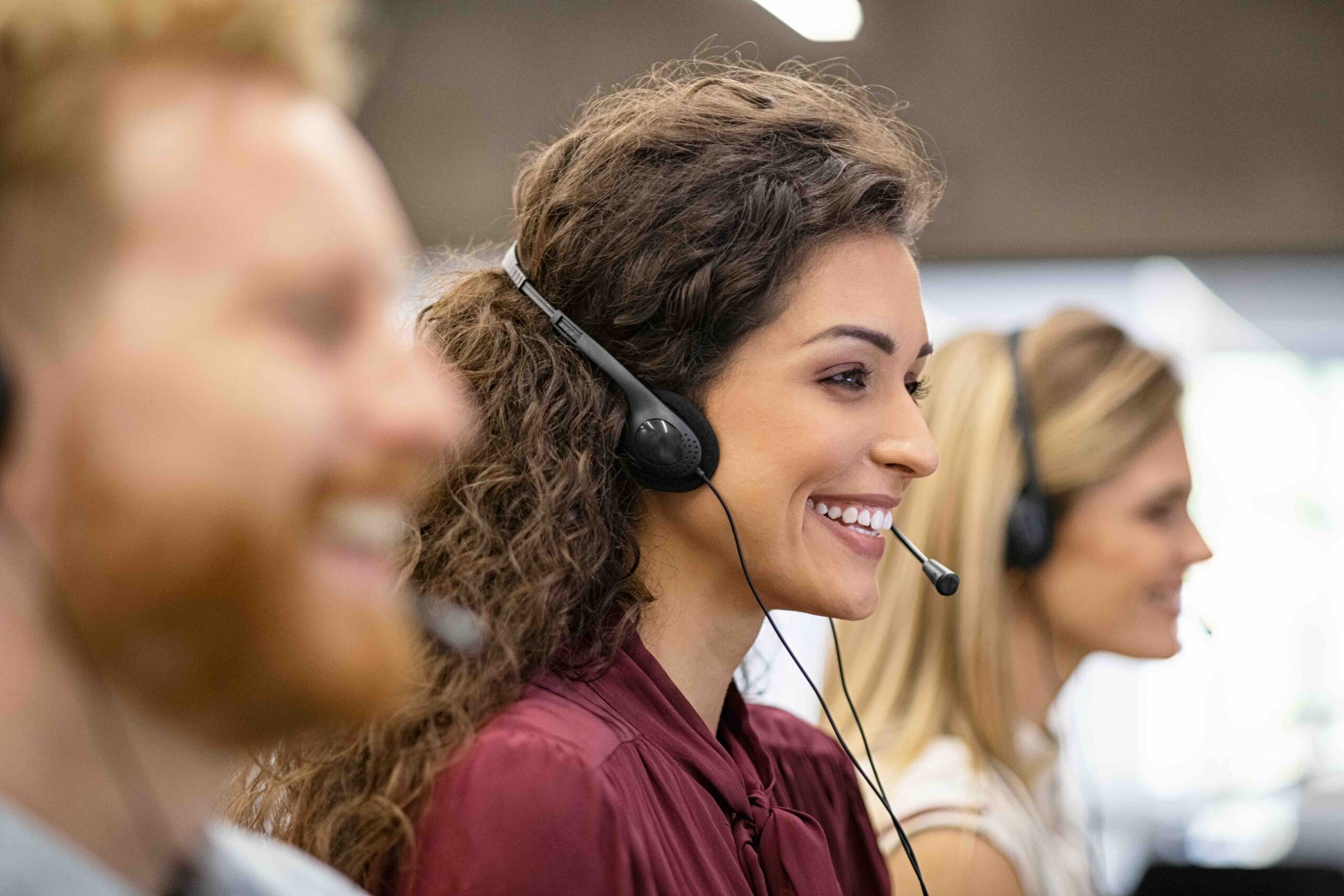 Smiling woman call center operator doing her job with a headset while working on computer. Positive smiling agents in conversation with customer over headset, sitting in row. Consulting and assistance helpdesk service.
